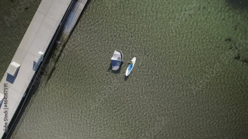Aerial view of a surfer sitting on his surfboard in the water next to a pier on a sunny day. 4K drone video.