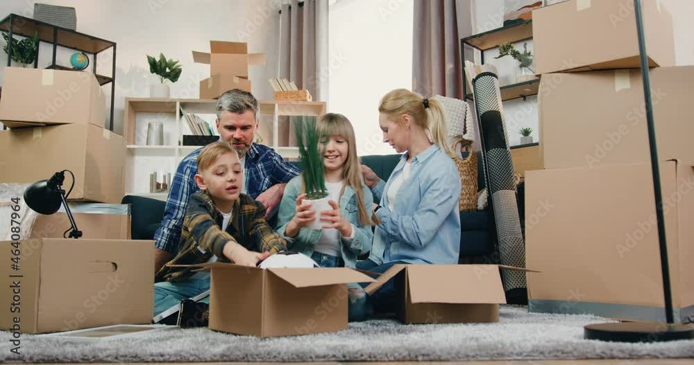 Likable smiling happy friendly modern family with two kids sitting on the floor among cardboard boxes in relocation day and unpacking different home decors Likable smiling happy friendly modern family with two kids sitting on the floor among cardboard boxes in relocation day and unpacking different home decors