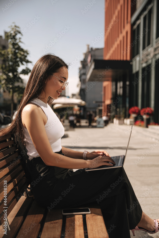 Profile photo of stylish asian student working on her thesis at laptop outdoors. Smiling brunette dressed in white top, black trousers.