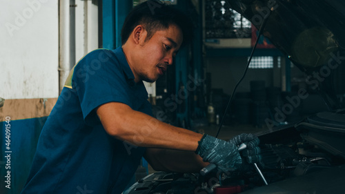 Professional car mechanic screwing details of car engine on lifted automobile at repair service station. Skillful Asian guy in uniform fixing car at mechanics garage at night.