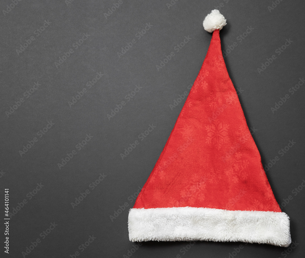 A red and white Santa hat with a snowflake pattern lies on the right against a dark matte background. Top view A red and white Santa hat with a snowflake pattern lies on the right against a dark matte background. Top view