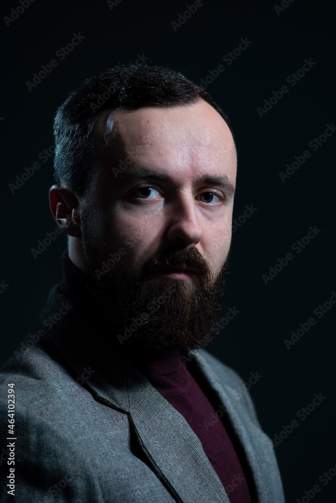 a dramatic portrait of a bearded millennial guy in a jacket and sweater. On a dark background a dramatic portrait of a bearded millennial guy in a jacket and sweater. On a dark background
