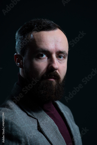 a dramatic portrait of a bearded millennial guy in a jacket and sweater. On a dark background