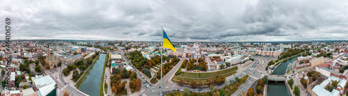 Flag of Ukraine on flagpole with epic heavy cloudscape, extremely wide city aerial panorama on autumn river Lopan embankment, Holy Annunciation Cathedral, central streets in Kharkiv, Ukraine