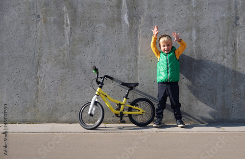Ecstatic funny little child boy standing near concrete wall with his yelllow bicycle on sunny day. Urban lifestyle.