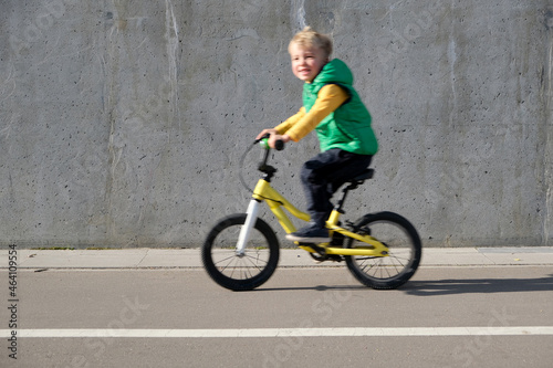 Happy little child boy in blurry focus is riding bicycle against static gray concrete wall. Cheerful kid cycling in the city in motion blur.