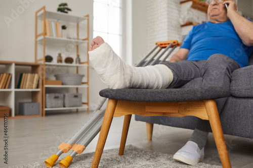 Senior man with broken leg in gypsum plaster cast sitting on sofa at home and talking on mobile phone. Elderly patient with injured foot resting on couch and calling doctor on cellphone. Close up