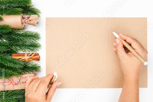 top view of woman hands writing New Year wish list on craft paper with frame made of Christmas tree branches and gift boxes. 