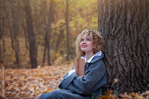 Beautiful girl reads a book in the autumn park. The girl has curly hair. Book in hand. Yellow autumn leaves. Copy space. Blurred background.
