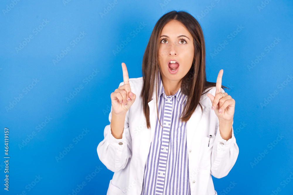 young european doctor girl wearing white medical gown on white background amazed and surprised looking up and pointing with fingers and raised arms. young european doctor girl wearing white medical gown on white background amazed and surprised looking up and pointing with fingers and raised arms.