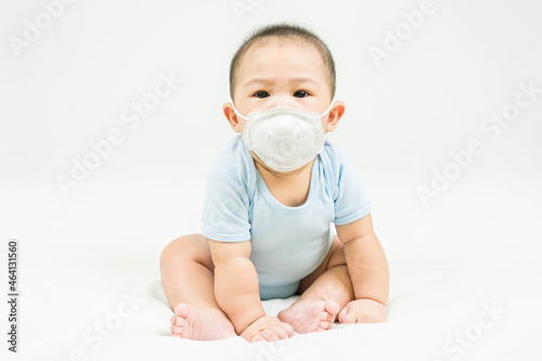 Concept covid-19 coronavirus pandemic.Portrait of cute little 5 months old asian newborn baby boy wearing medical mask in blue bodysuit sitting on white blanket at home protect from infection of virus