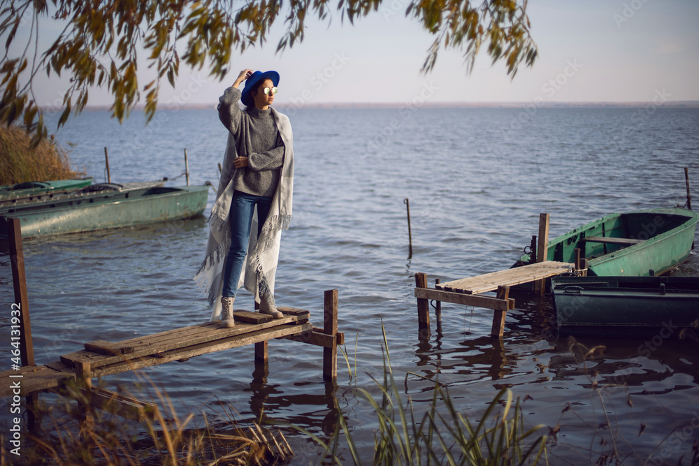 woman in jeans and a sweater and a hat stands on a pier with boats by lake in autumn