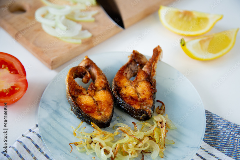 Fried fish with onions is on a plate, on a waffle towel, next to it is a cutting board with chopped onions, lemon and tomato on a white background. High quality photo Fried fish with onions is on a plate, on a waffle towel, next to it is a cutting board with chopped onions, lemon and tomato on a white background. High quality photo