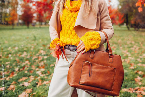 Close up of stylish woman holding purse wearing yellow sweater in autumn park. Fall female clothes  accessories. Fashion