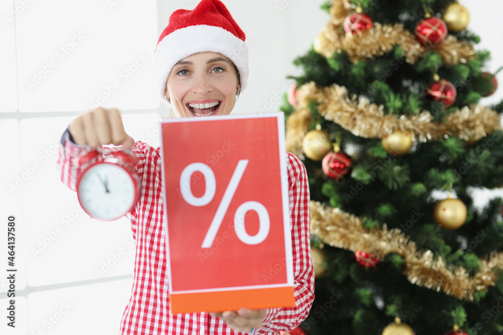 Young smiling woman in santa claus hat holds alarm clock and poster with percent on background of New Year tree Young smiling woman in santa claus hat holds alarm clock and poster with percent on background of New Year tree