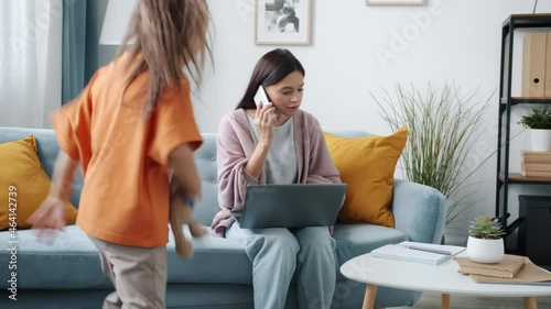 Busy young woman is working with laptop and making phone call when annoying child is demanding mother's attention. Childhood and remote work concept.