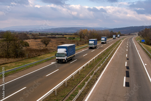 Highway transportation scene with Caravan or Convoy of Blue transportation trucks in line on a rural highway under a dramatic sky.