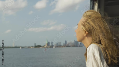 A young teenage girl takes in the view of lower Manhattan from a ferry. Blond woman watching liberty statue on background from the pier in the harbor. Travel, adventure, tourism concept.