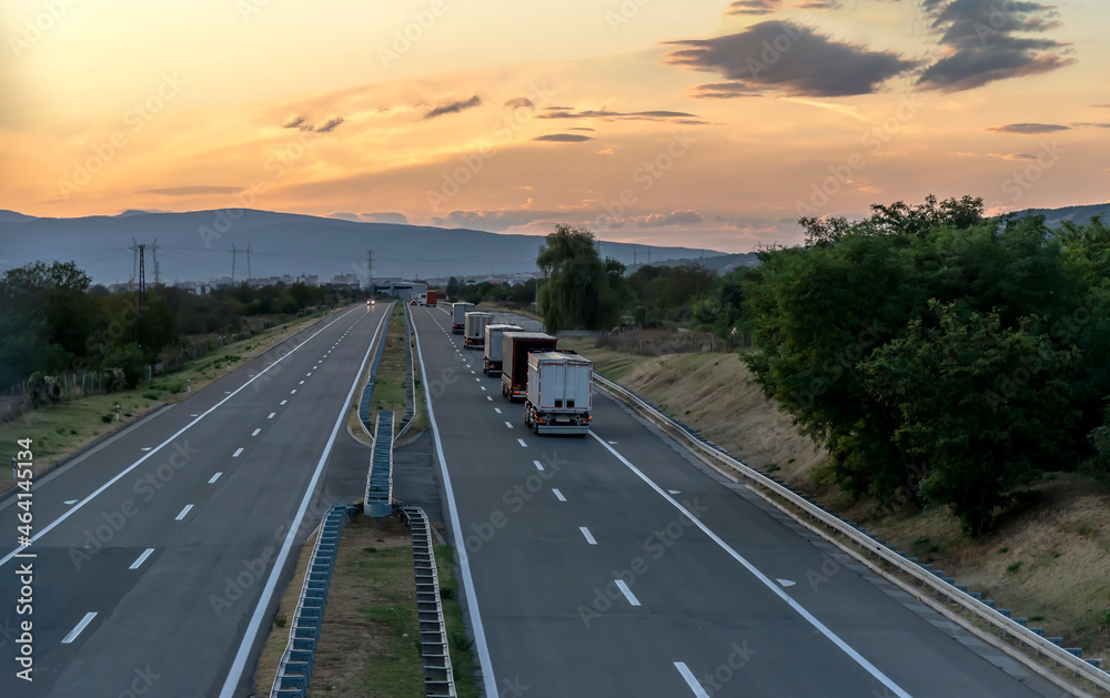 Convoy of Big Transportation trucks departing on a country highway under a sunset sky. 