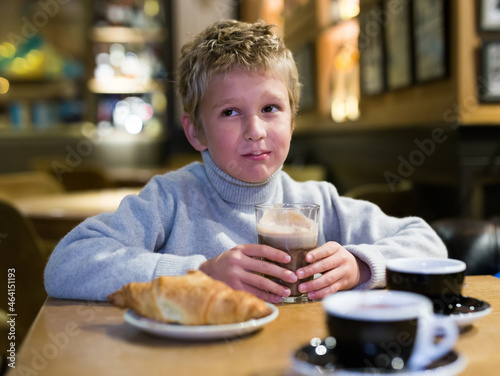 Portrait of happy cute preteen boy enjoying hot sweet chocolate with fresh croissant in cozy cafe..