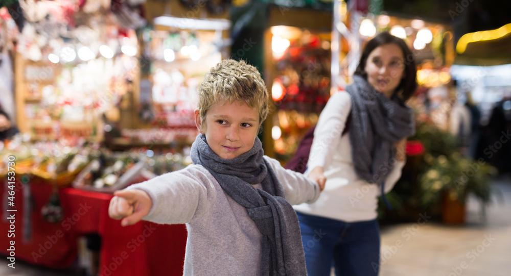 Portrait of tween boy pointing to desired thing and pulling mothers hand during walk at festive fair on Christmas Eve