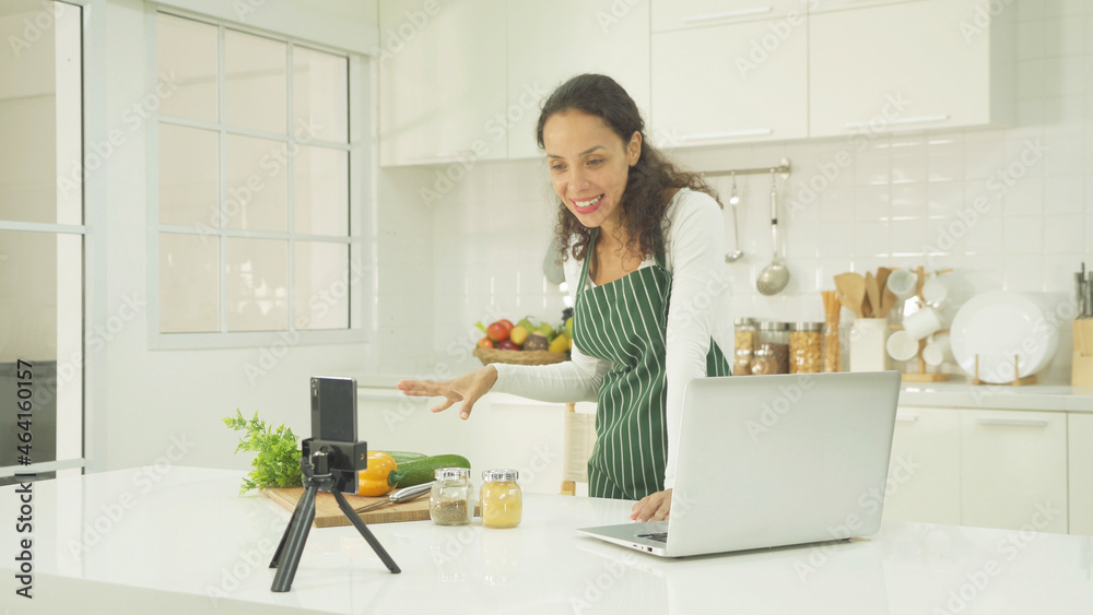 Portrait of happy smiling, latin woman talking, cooking food at home, video live on webcam video meeting with computer laptop, call conference, working online in kitchen. People lifestyle. Webinar