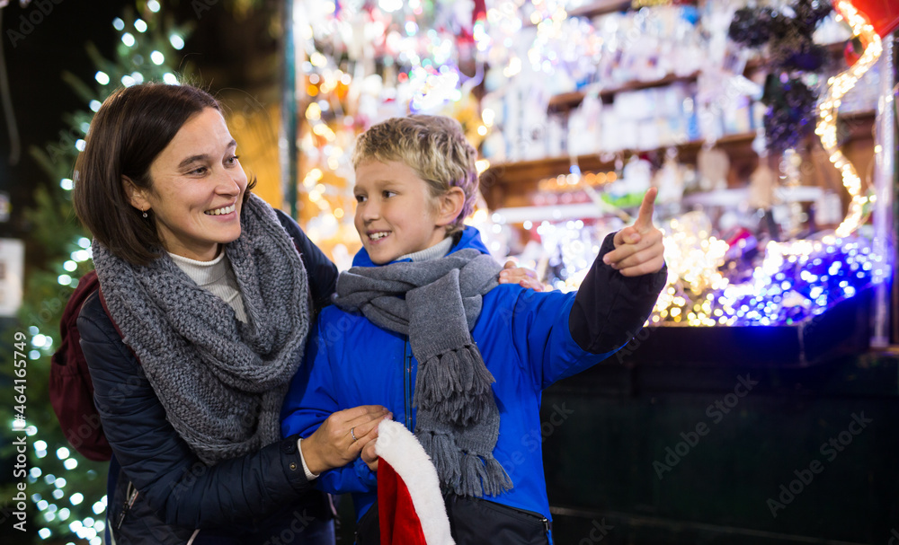Obraz premium Happy tween boy choosing Xmas decorations and gifts on street fair with his loving mother..