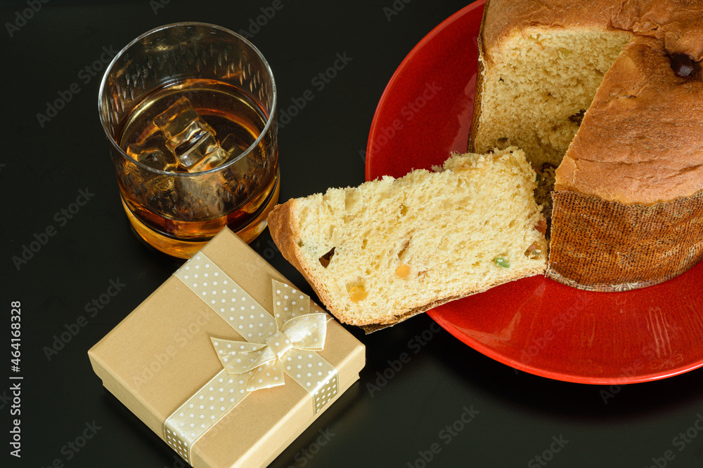 Fototapeta Slice of panettone next to a glass of whiskey and ice and a golden gift box.