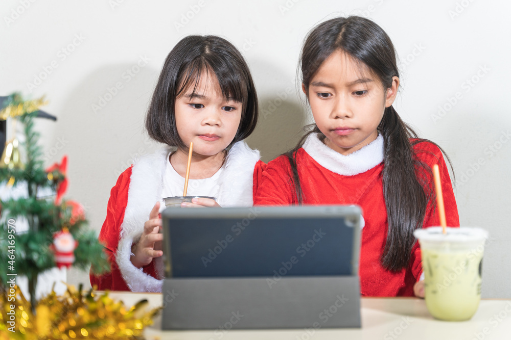 Two Asian children little girls wear Christmas Santa Claus chating with friend on digital tablet. Christmas Holiday Concept Stock Photo Two Asian children little girls wear Christmas Santa Claus chating with friend on digital tablet. Christmas Holiday Concept Stock Photo