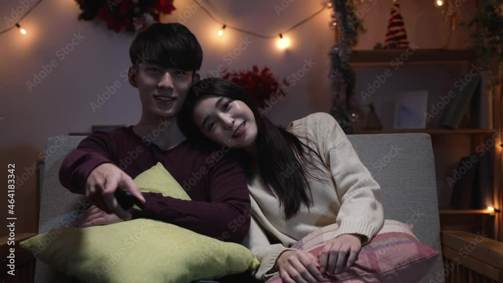 sweet asian young couple in love face camera at Christmas eve enjoy watch tv while sitting under pillows on sofa in living room. korean date partner switch channel by remote control on xmas evening sweet asian young couple in love face camera at Christmas eve enjoy watch tv while sitting under pillows on sofa in living room. korean date partner switch channel by remote control on xmas evening