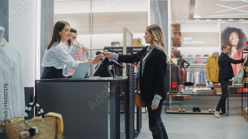 Clothing Store Checkout Cashier Counter: Beautiful Young Woman Buys Blouse from Friendly Retail Sales assistant, Paying with Contactless Credit Card. Fashion Shop with of Designer Brands.