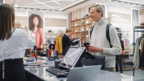 Clothing Store: Young Man At Checkout Counter Buys Clothes Paying with Contactless NFC Smartphone, Friendly Retail Sales Assistan Packs Merchandise. Contemporary Fashion Shop with of Designer Brand