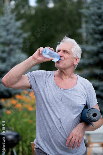 A handsome athletic fit gray-haired man drinks cool water from a bottle after a yoga class in the backyard. He holds a yoga mat in his hand