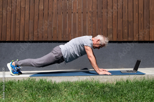 A fit athletic elderly man does yoga on the background of his house using a laptop
