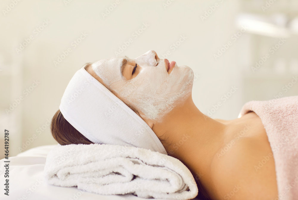Professional cosmetology and treatment. Beautiful woman with headband and cleansing white mask on her face relaxes in spa or beauty salon. Side view close up of woman with closed eyes lying on towel.