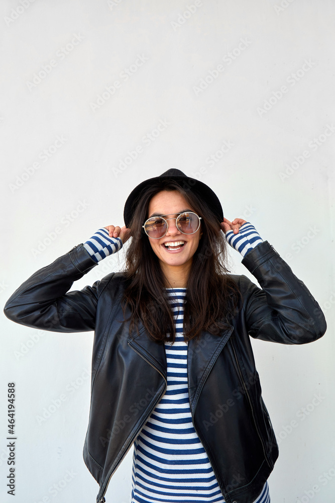 Stylish woman in leather jacket and hat. Delighted trendy female in black leather jacket and sunglasses touching hat and looking at camera on white background Stylish woman in leather jacket and hat. Delighted trendy female in black leather jacket and sunglasses touching hat and looking at camera on white background