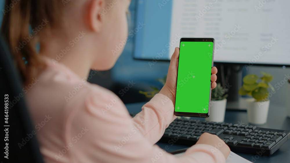 Naklejka premium Close up of child holding green screen on smartphone for homework and school tasks. Young girl looking at display with mockup template and isolated background for remote education.