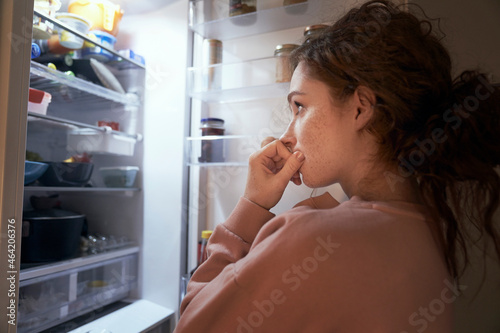Fototapeta Undecided young caucasian woman checking fridge for some food at night