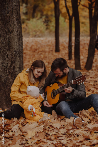 Happy young family sitting with little baby at the autumn park, dad plays on guitar