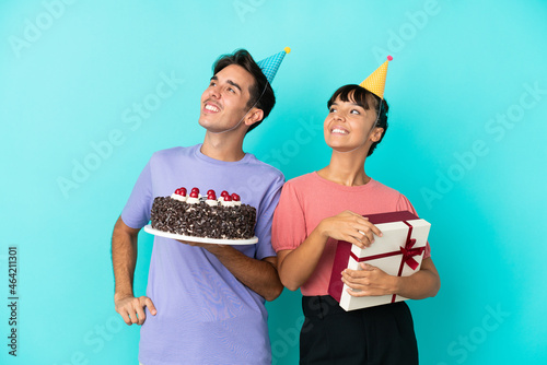 Young mixed race couple holding birthday cake and present isolated on blue background looking up while smiling