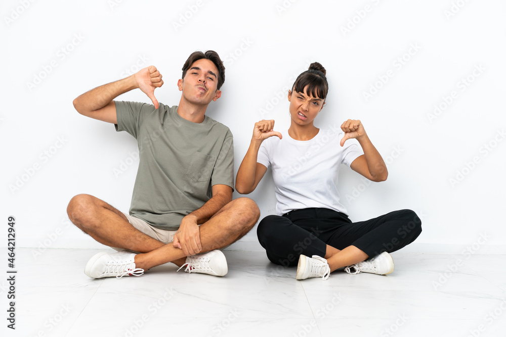 Young mixed race couple sitting on the floor isolated on white background showing thumb down with both hands Young mixed race couple sitting on the floor isolated on white background showing thumb down with both hands