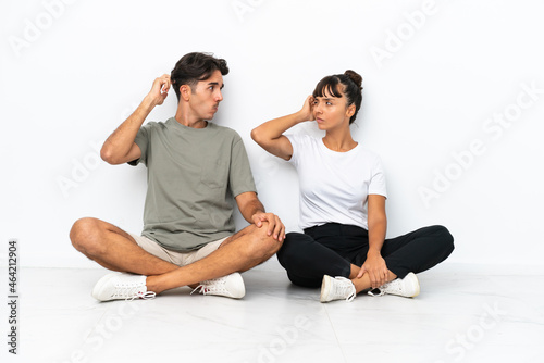 Young mixed race couple sitting on the floor isolated on white background having doubts while scratching head