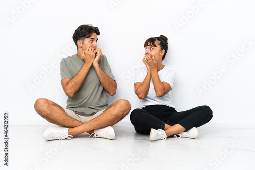 Young mixed race couple sitting on the floor isolated on white background is a little bit nervous and scared putting hands to mouth
