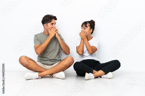 Young mixed race couple sitting on the floor isolated on white background covering mouth with hands for saying something inappropriate