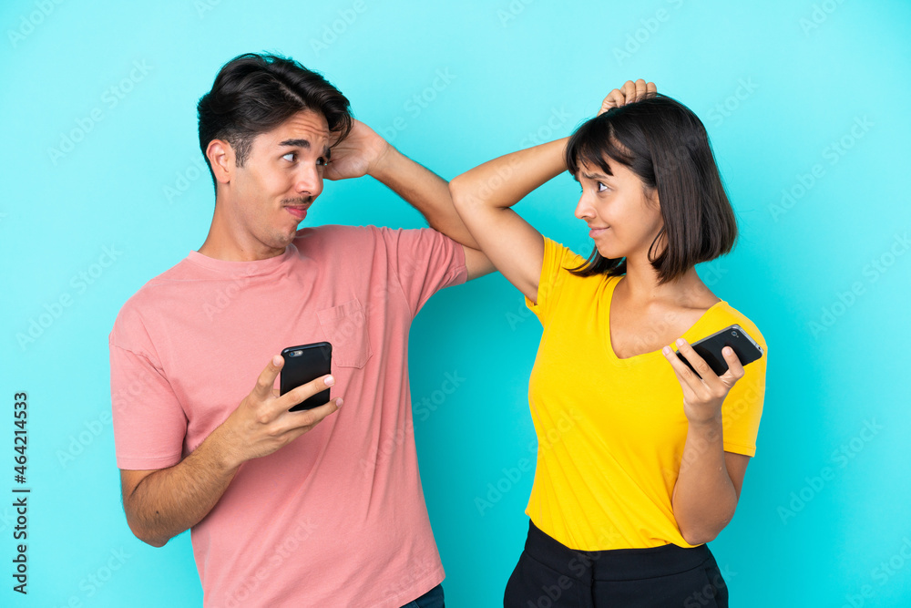 Young mixed race couple holding mobile phone isolated on blue background having doubts while scratching head Young mixed race couple holding mobile phone isolated on blue background having doubts while scratching head