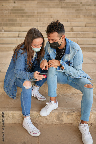 Friends with face masks sitting on stairs outdoors looking and pointing at the smart phone