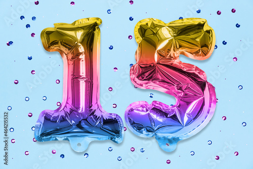 Rainbow foil balloon number, digit fifteen on a blue background with sequins. Birthday greeting card with inscription 15. Anniversary concept. Top view. Numerical digit. Celebration event, template.