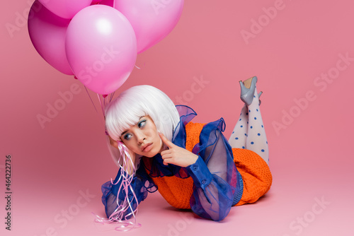 Pensive asian pop art woman holding balloons while lying on pink background