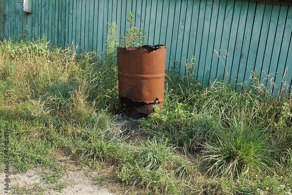Obraz premium one red old rusty iron barrel with a large hole stands in the green grass near the wooden wall of the fence on the street