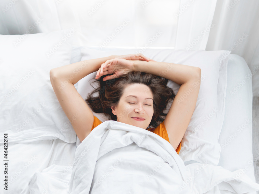Woman stretches while lying in bed. Pleasant awakening after night's sleep. White bed linen. Top view.